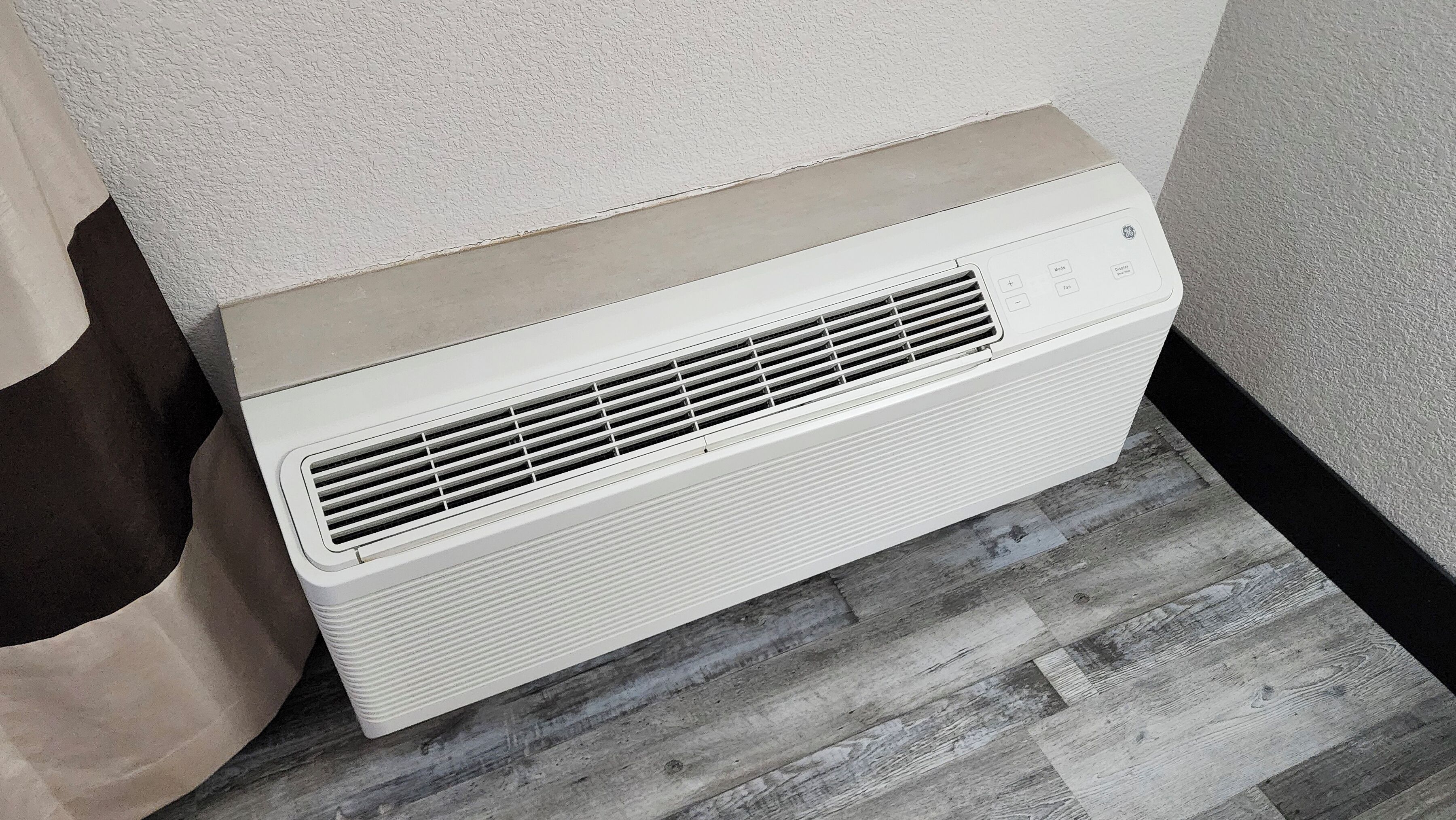 A white portable air conditioner against a textured wall and wooden floor.