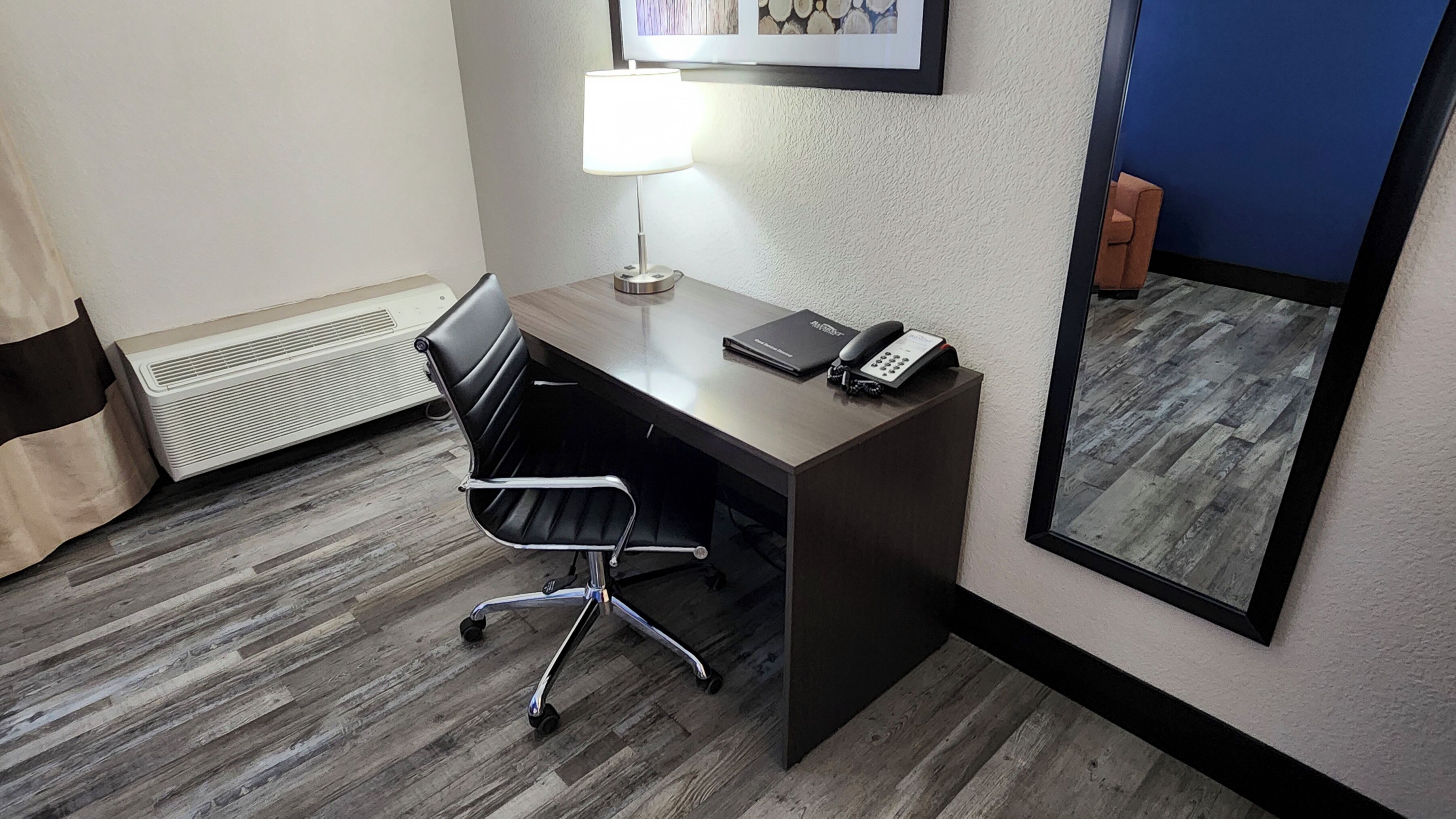 A hotel room with a desk, chair, lamp, phone, and a large mirror.