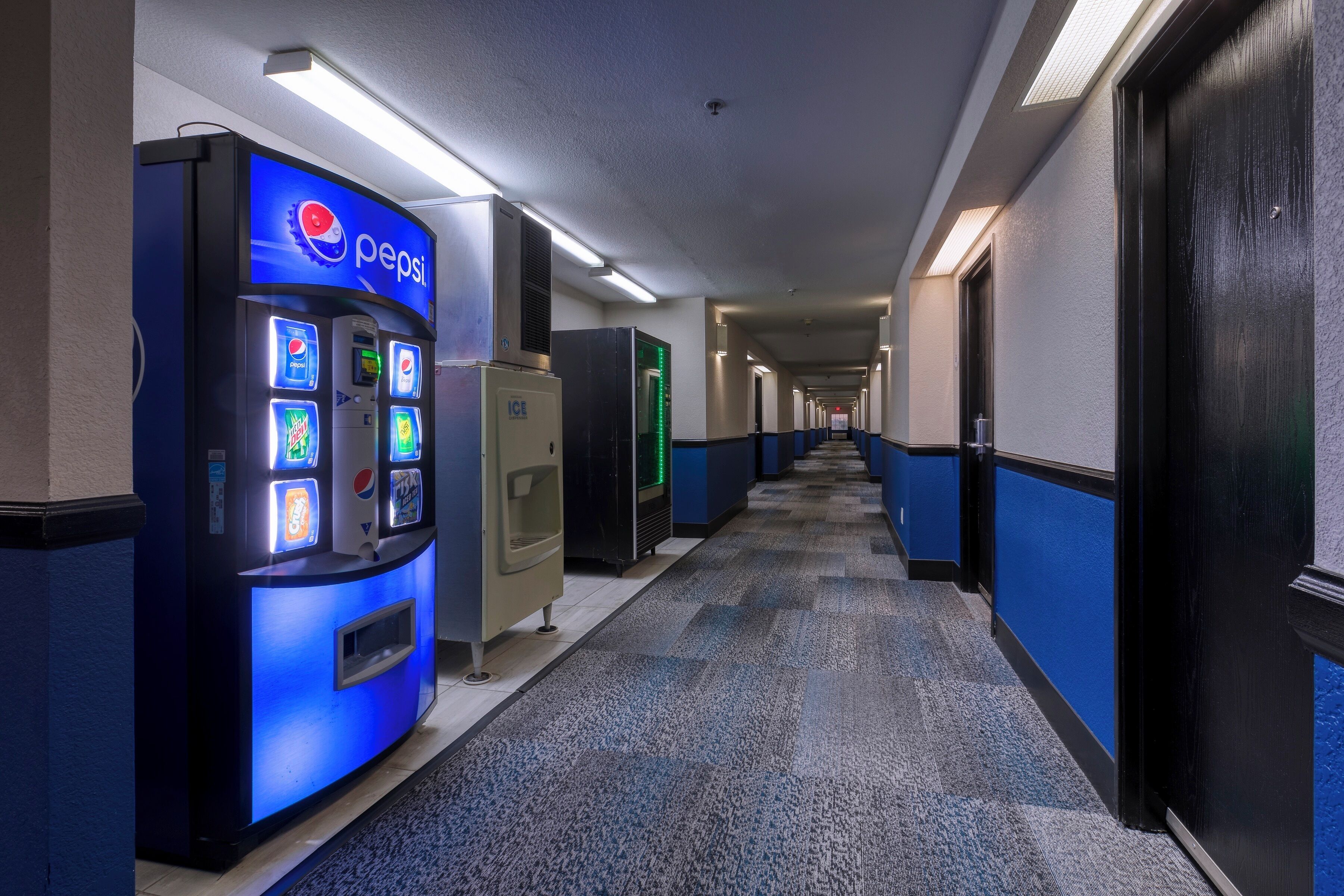 Corredor de hotel com máquina de Pepsi, bebedouro e frigorífico iluminado a verde.