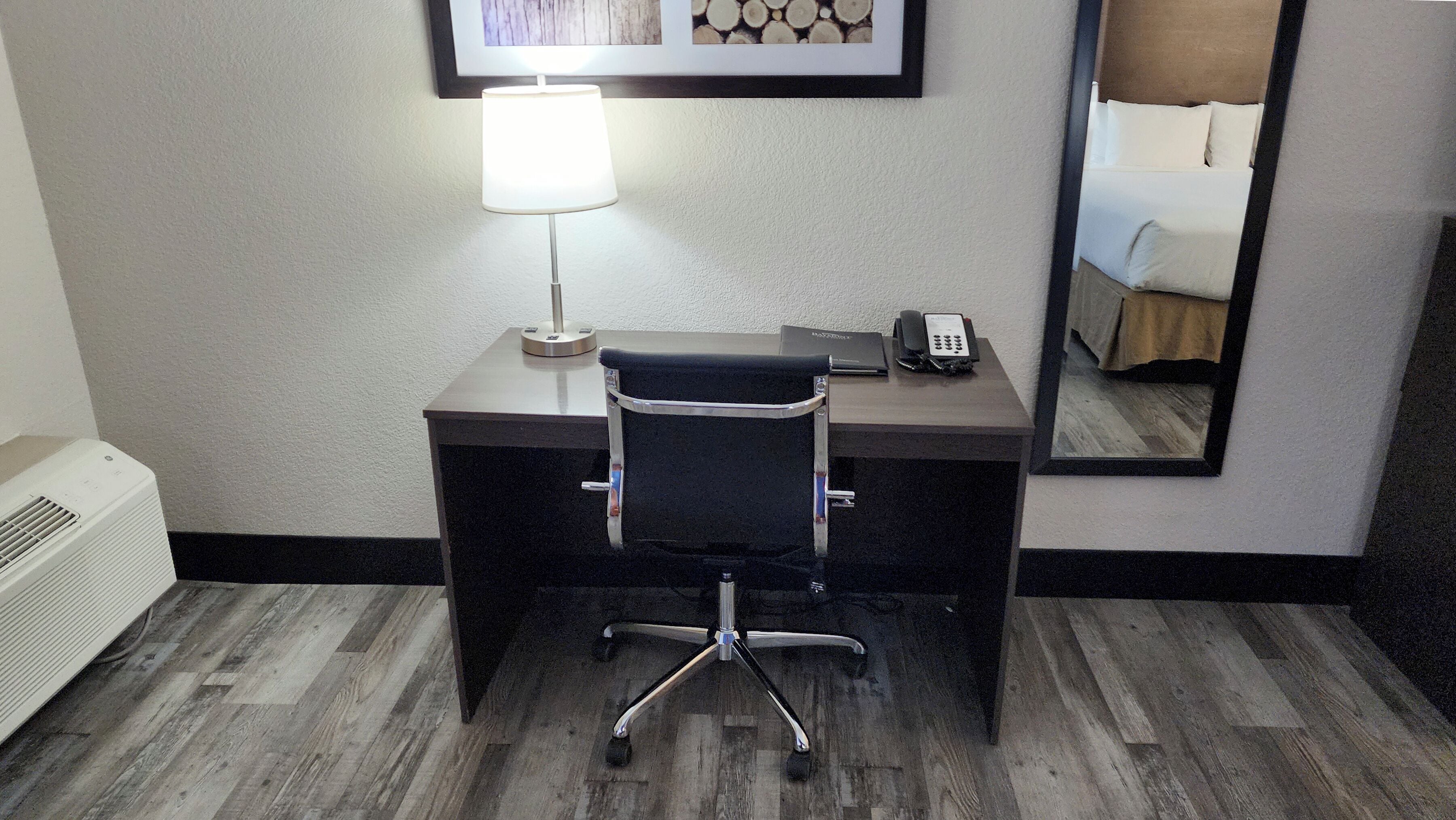 A hotel room with a desk, chair, lamp, phone, and a mirror reflecting a bed.