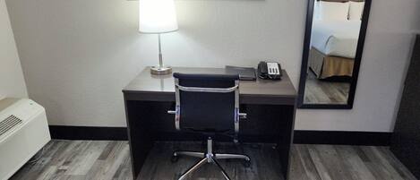 Habitación de hotel con escritorio, silla, lámpara, teléfono y un espejo que refleja una cama.