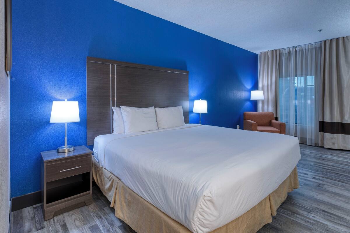 Ein Hotelzimmer mit einem großen Bett, zwei Nachttischlampen, einem Nachttisch, einem Sessel und einem Fenster mit Vorhängen.