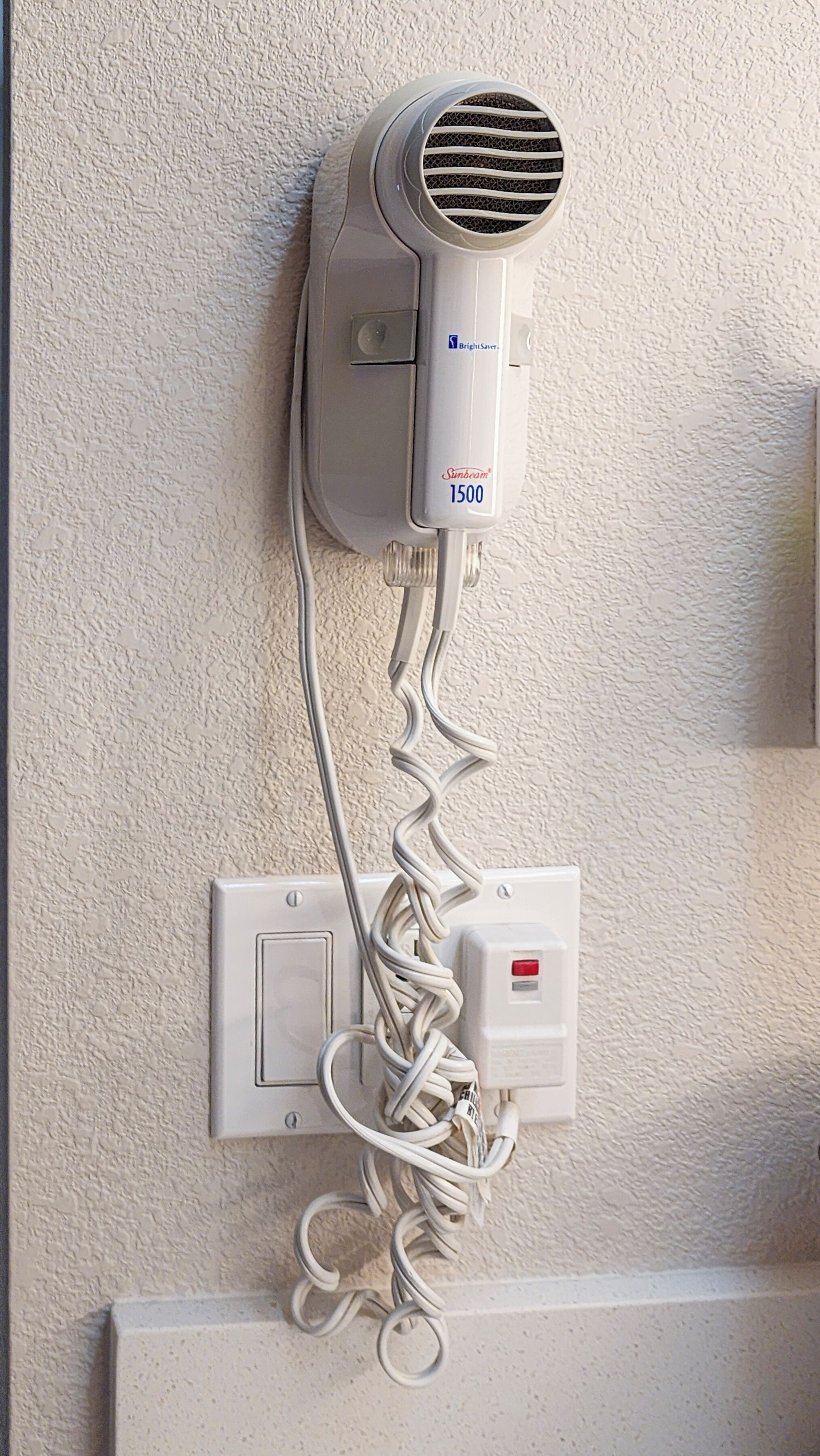 A hair dryer mounted on a textured wall, connected to a power outlet with a coiled cord.