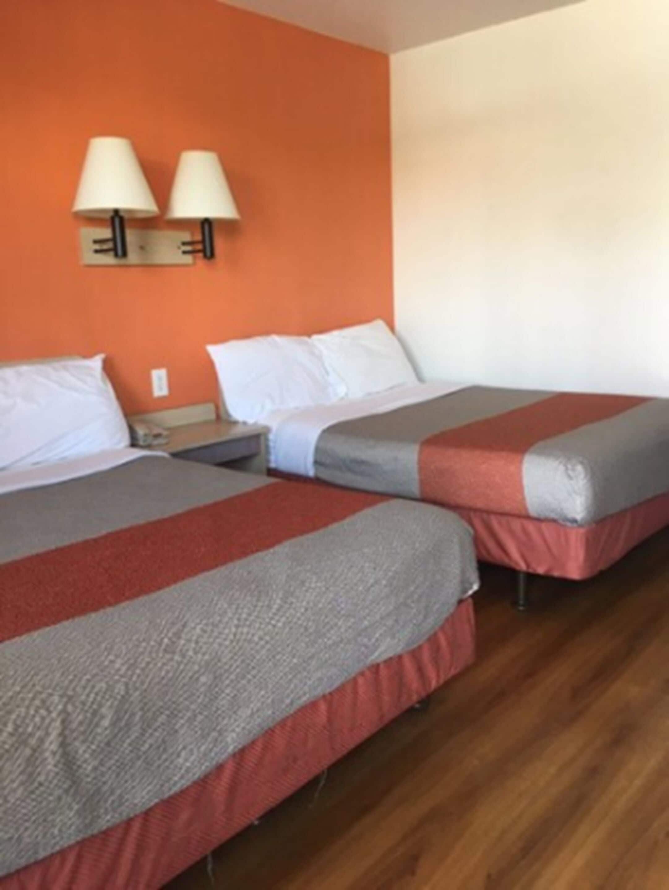 A hotel room with two beds, each with a red and gray blanket, a nightstand, a lamp, and an orange wall.