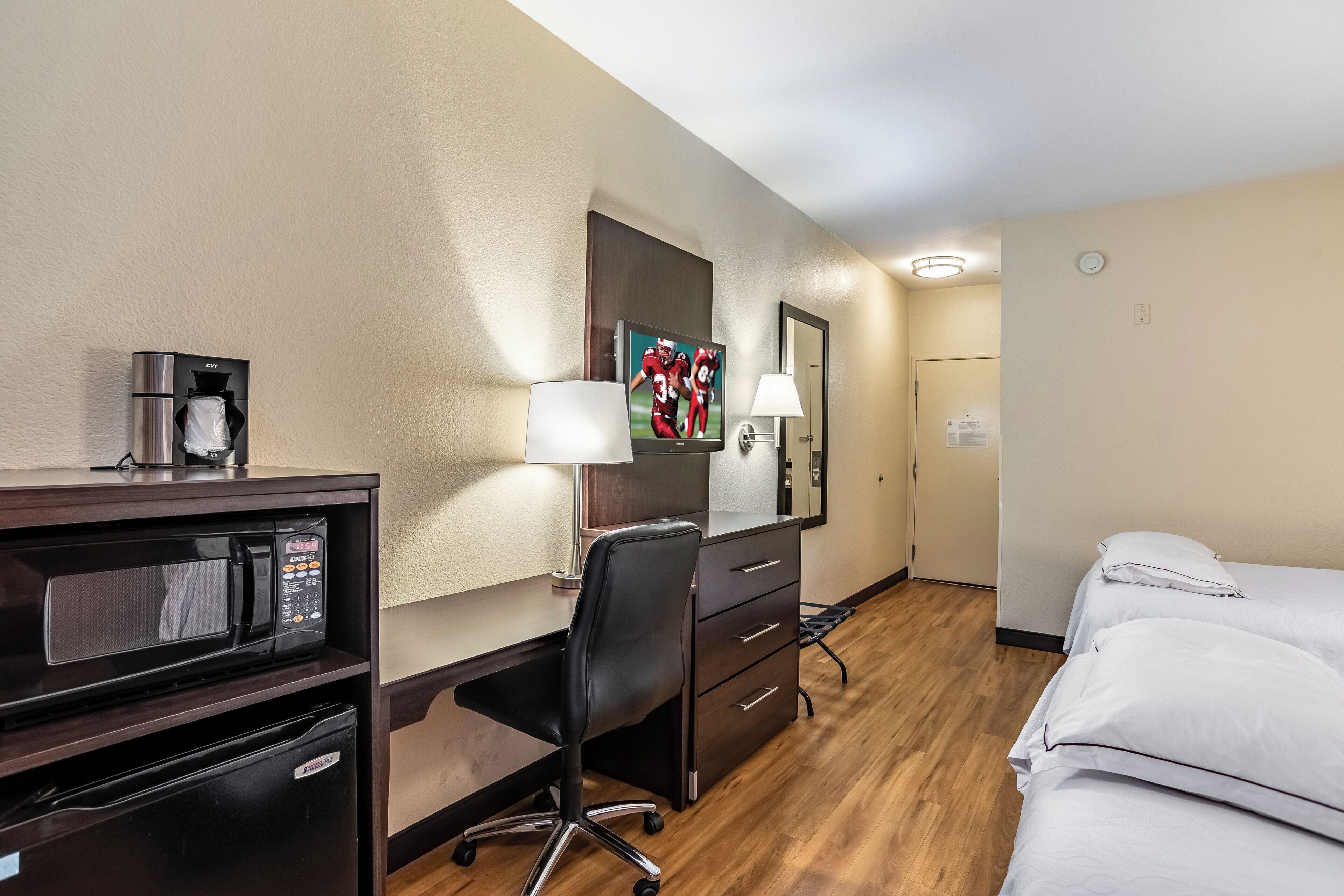 A hotel room with a desk, a chair, a bed, a microwave, a coffee maker, and a TV.