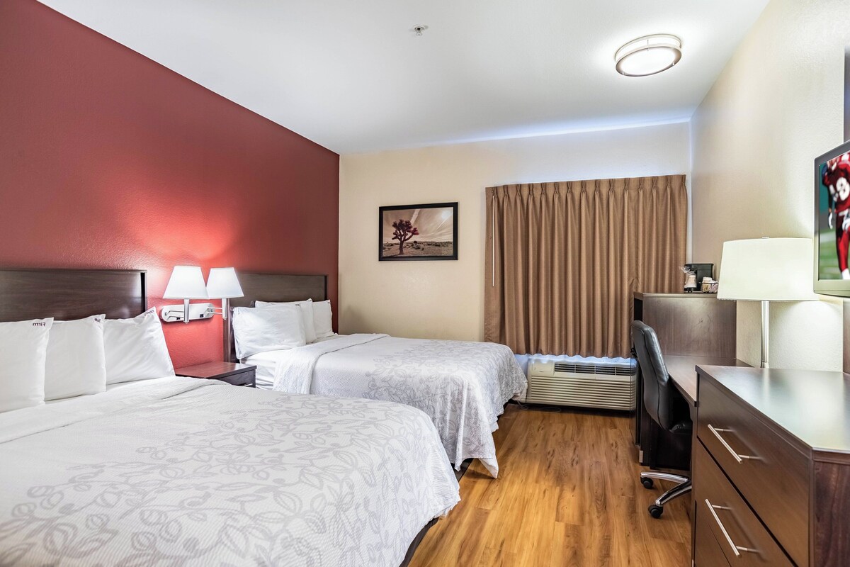 Ein Hotelzimmer mit zwei Betten, einem Schreibtisch, einem Fernseher und einem Vorhangfenster.
