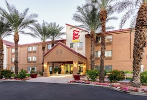 A Red Roof Inn with a traditional architectural style, featuring a central entrance with a covered walkway, surrounded by palm trees and flower beds.