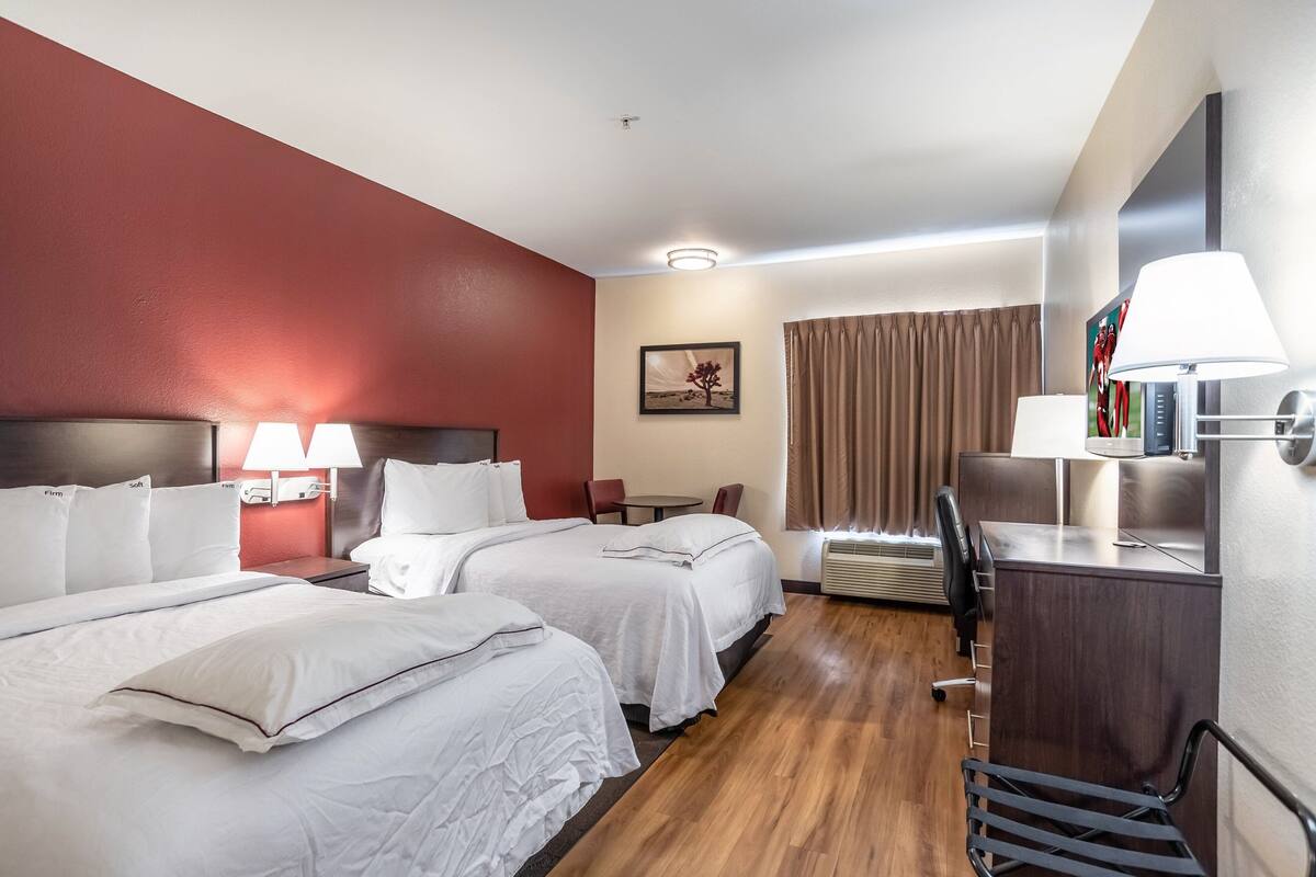 Una habitación de hotel con dos camas, un escritorio, una silla, una lámpara y un cuadro en la pared.