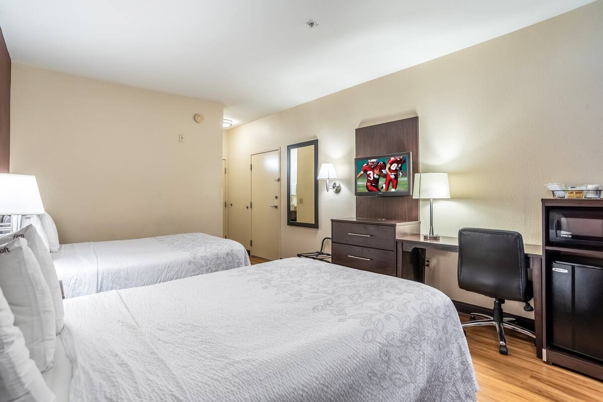 Quarto de hotel com duas camas, uma mesa de escritório com luminária, televisão e um pequeno espaço para trabalho.