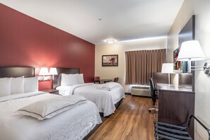 Ein Hotelzimmer mit zwei Betten, einem Schreibtisch, einem Stuhl, einer Schreibtischlampe, einem Bild an der Wand und einem Fenster mit Vorhängen.