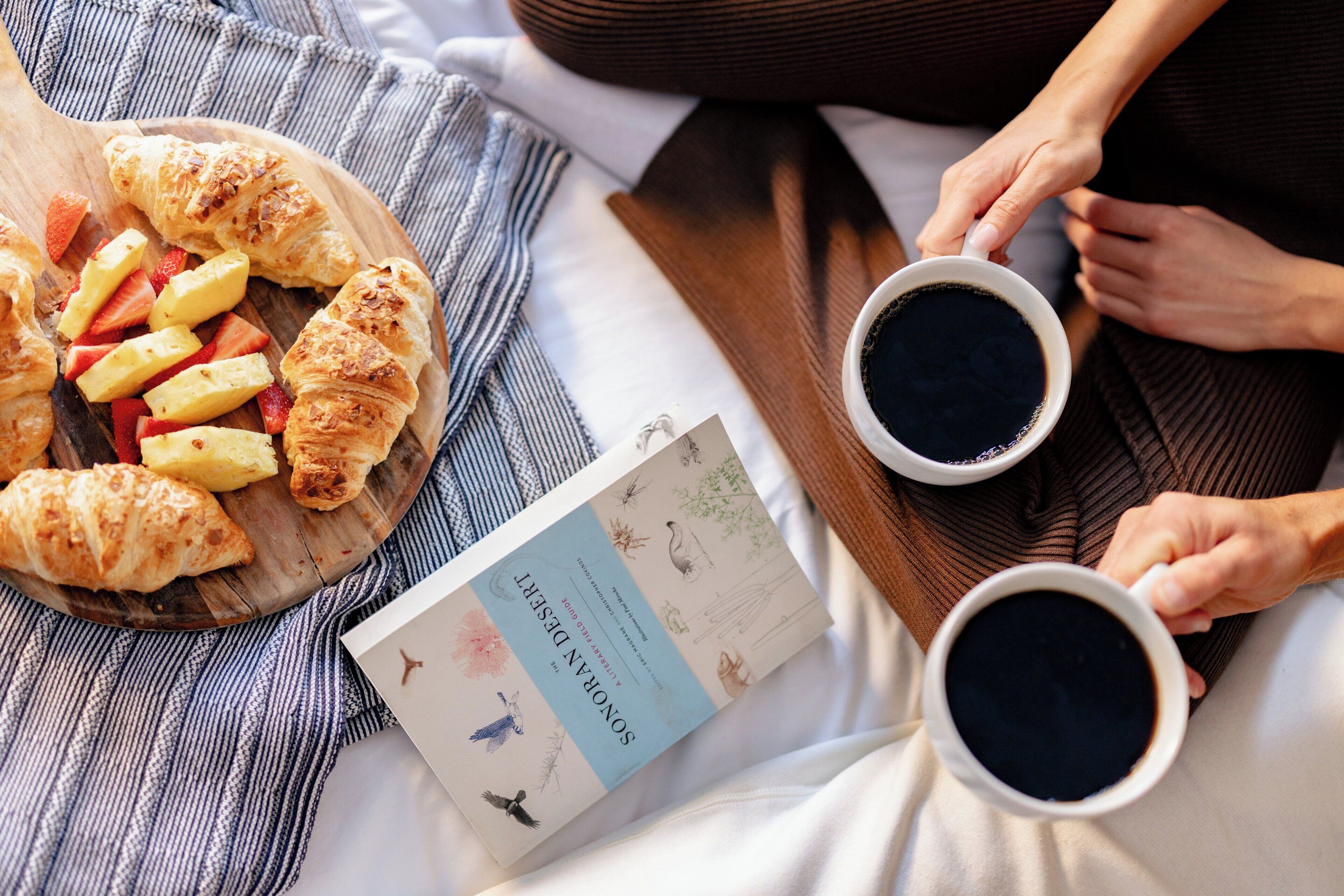 A breakfast setup with croissants, strawberries, and a book on a bed.