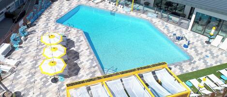 A swimming pool with sun loungers and umbrellas, surrounded by a checkered floor and a building with large windows.