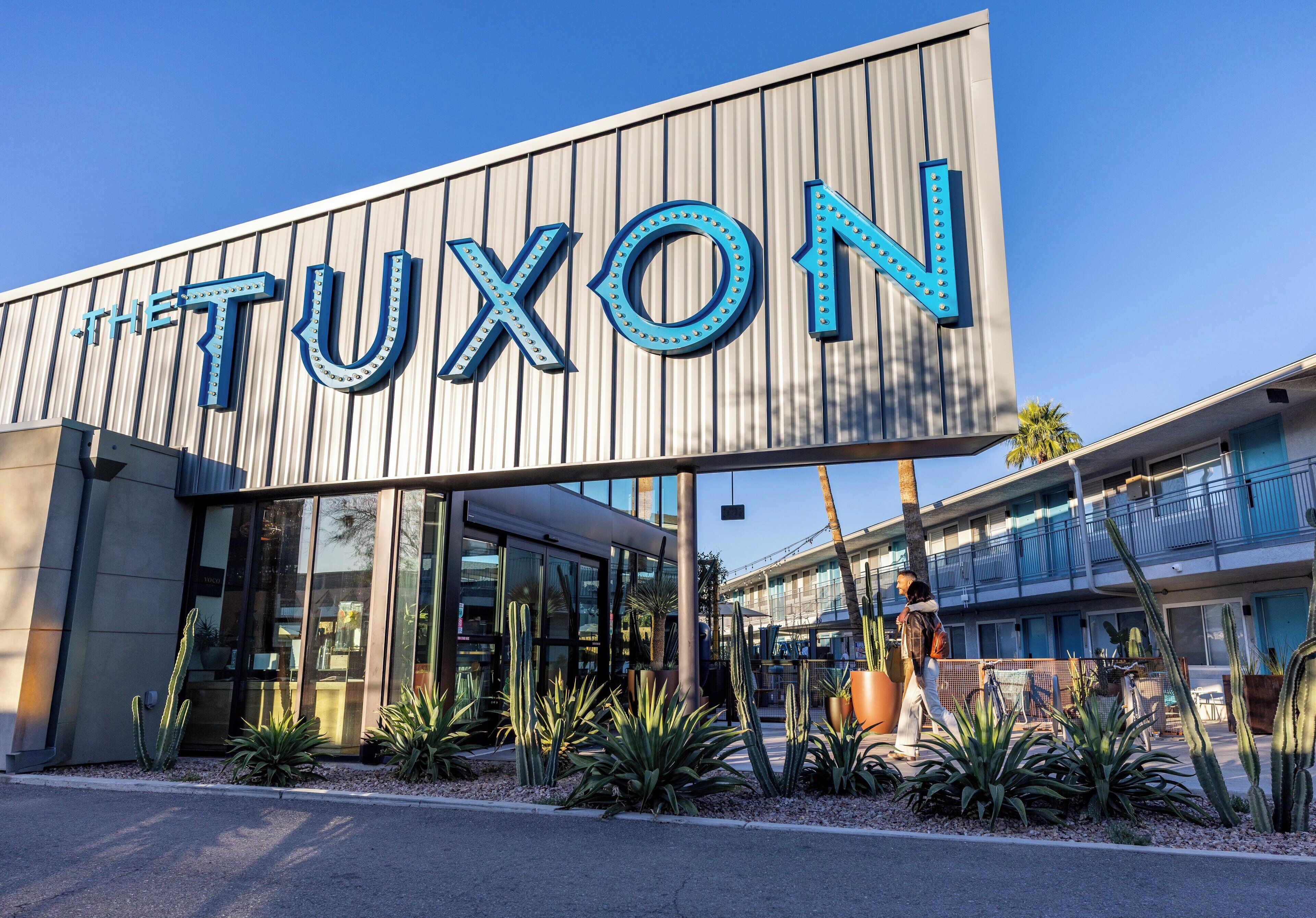 A building with a sign reading "THE TUXON" and a modern architectural design with large windows and a landscaped area with cacti.