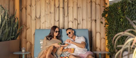 A couple sitting on a bed with a wooden backdrop, eating a meal and drinking.