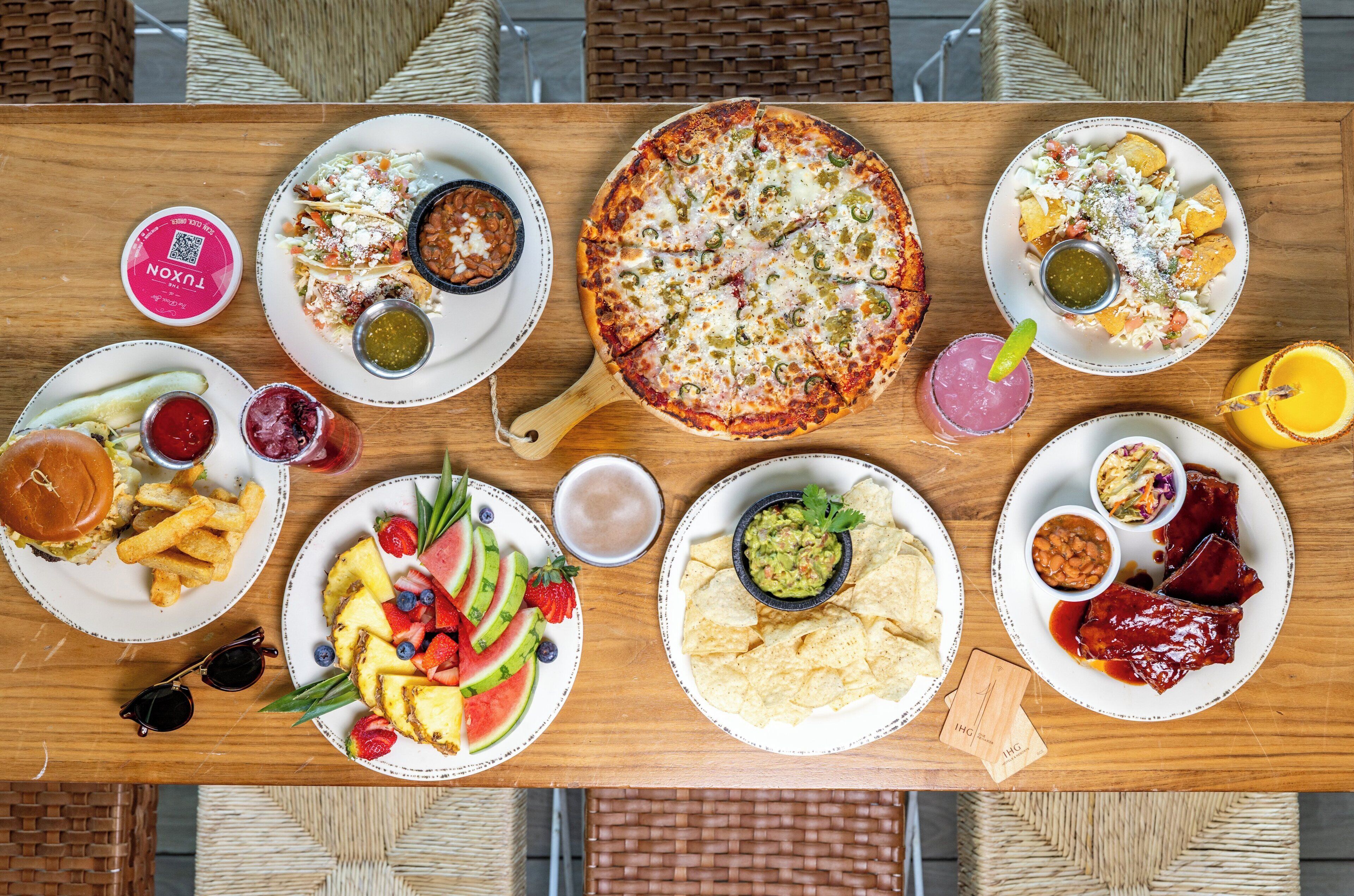 Et træbord med forskellige retter, herunder pizza, nachos og en burger.