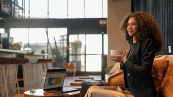 Una persona sentada en un sofá de cuero en una cafetería moderna, con una computadora portátil sobre una pequeña mesa redonda y sosteniendo una taza.