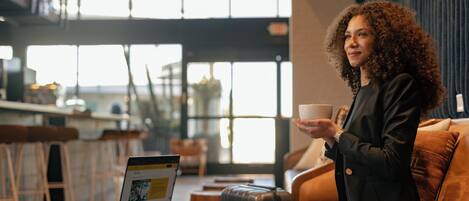 A person sitting on a leather couch in a modern cafe with a laptop on a small round table, holding a cup.