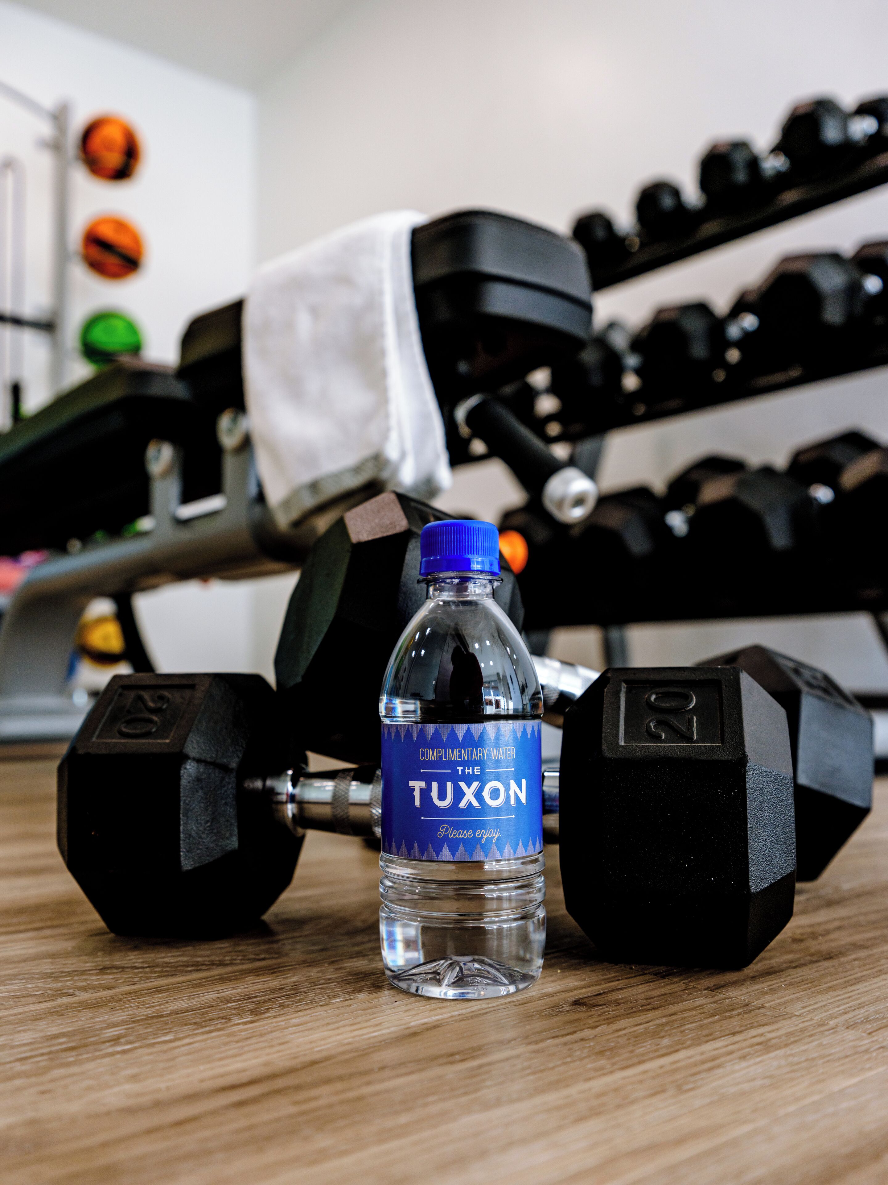 Una botella de agua con la etiqueta 'Tuxon' y dos mancuernas sobre una superficie de madera.