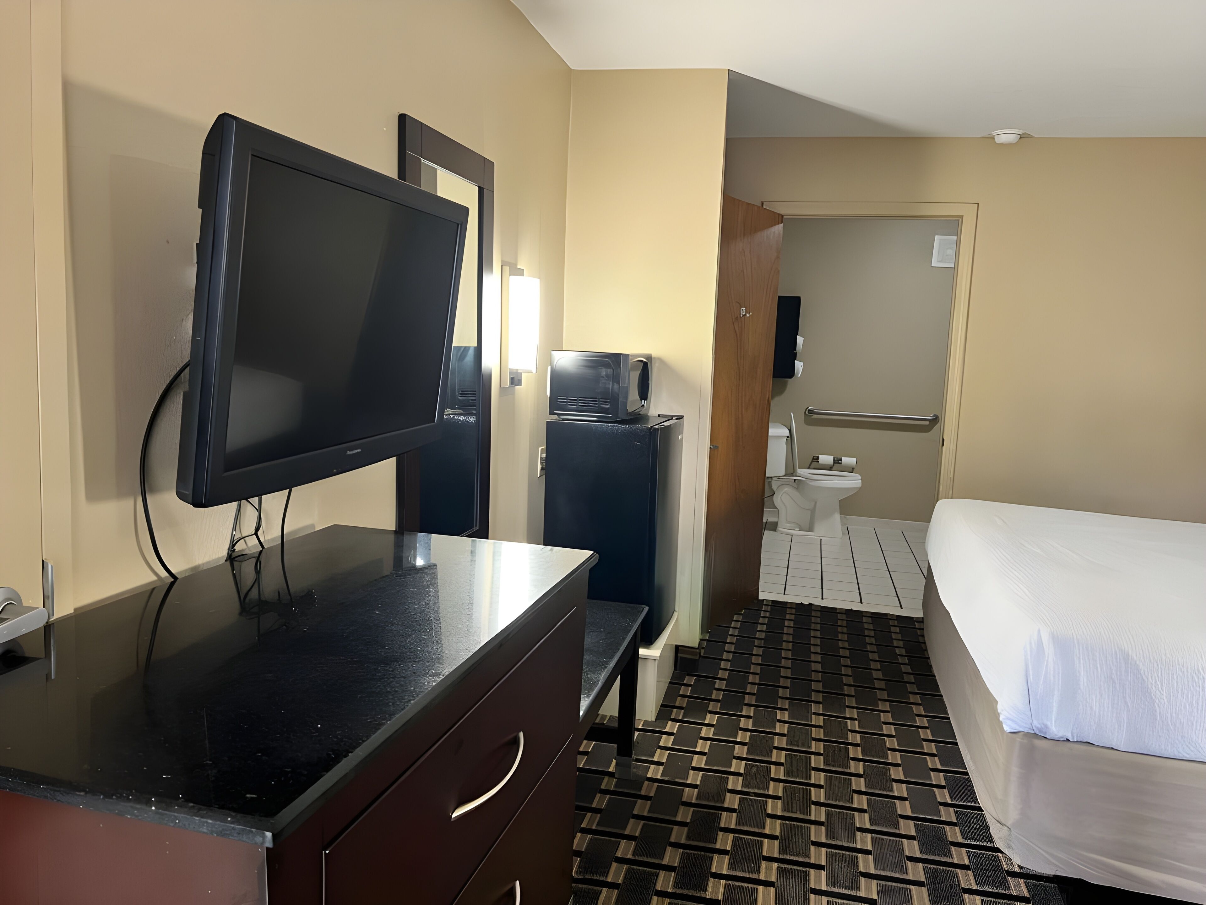 A hotel room with a bed, a TV, a desk with drawers, a small refrigerator, and a bathroom with a toilet and sink.