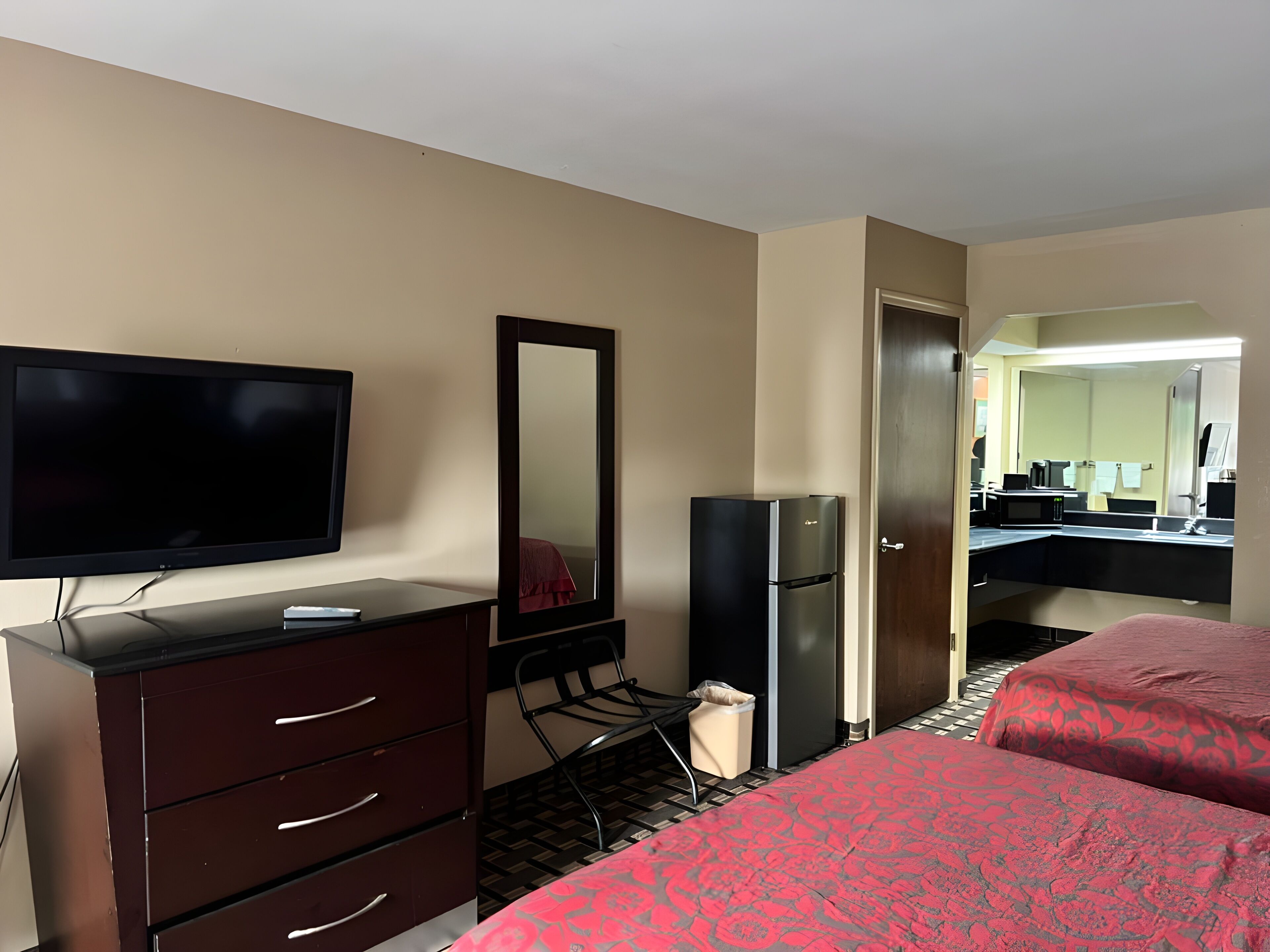 A hotel room with a TV, a mirror, a refrigerator, a dresser, and two beds with red bedding.