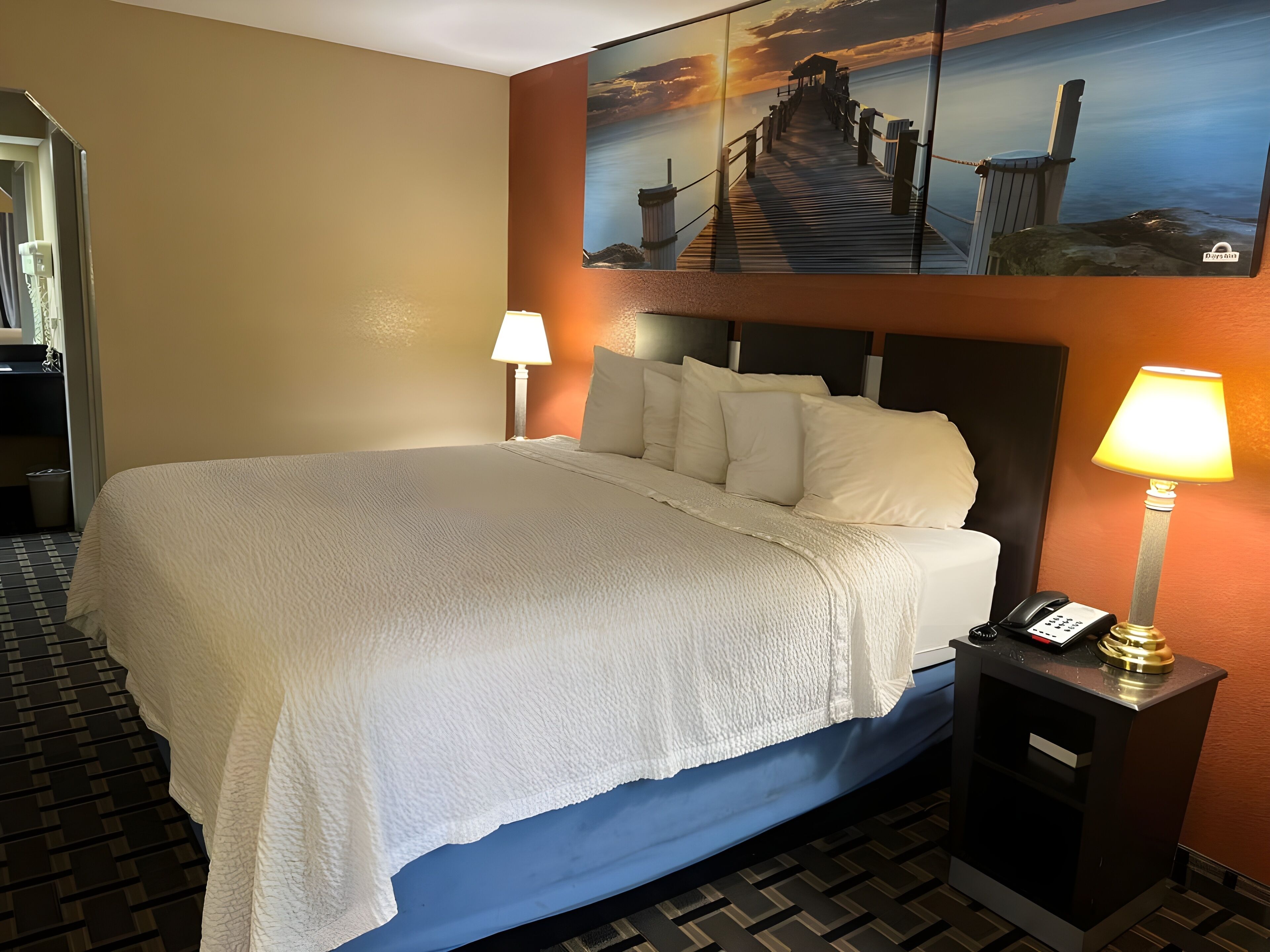 A hotel room with a large bed, a nightstand with a lamp, a painting of a pier, and a telephone.