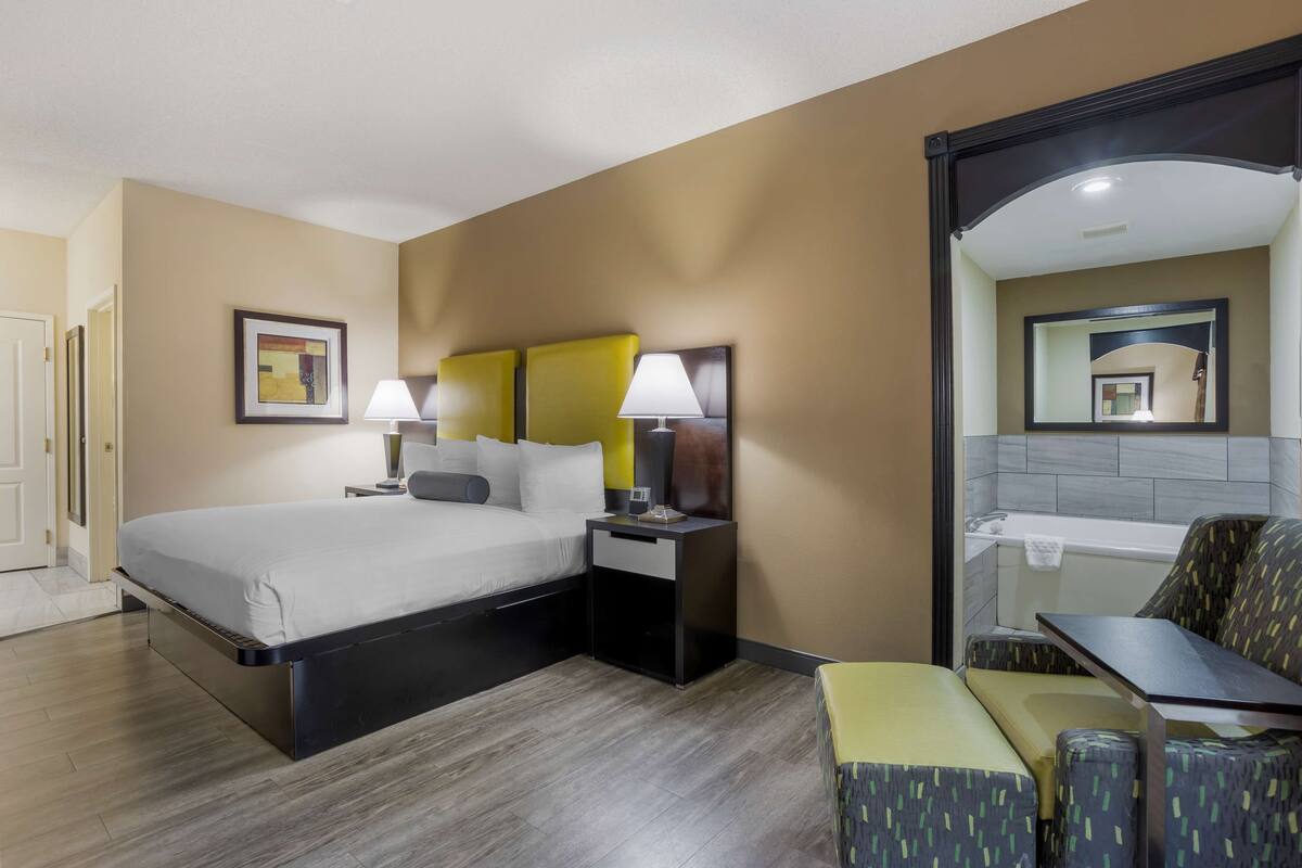 A hotel room with a bed, bedside tables, a chair, a small table, and a bathroom mirror.