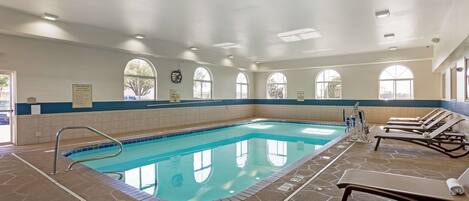 An indoor swimming pool with clear blue water, surrounded by stone tiles and equipped with lounge chairs and a pool ladder.
