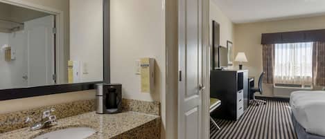 A hotel room with a granite countertop sink, a coffee maker, a desk with a chair, a bed, and a window with curtains.