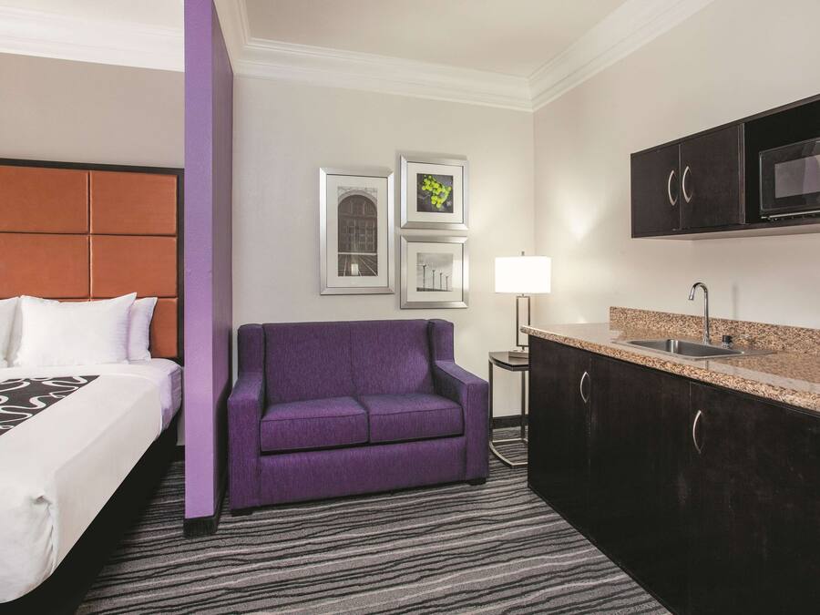 Ein Hotelzimmer mit einem Bett, einem violetten Sessel, einem kleinen Tisch mit Lampe und einer Küchenzeile mit Mikrowelle.