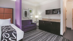 Habitación de hotel con una cama, un sofá morado, un escritorio pequeño con lámpara, una kitchenette con microondas y cuadros en la pared.