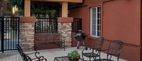 An outdoor patio area with a stone wall, a wooden pergola with string lights, a grill, and metal chairs and tables.