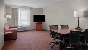 Una habitación de hotel con alfombra roja, un televisor sobre un mueble de madera, una mesa de conferencias con sillas, un sofá y una lámpara.
