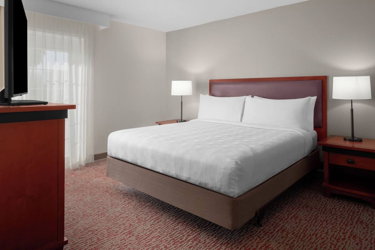 Een hotelkamer met een groot bed, twee nachtkastjes met lampen, een televisie op een standaard en een raam met dunne gordijnen.