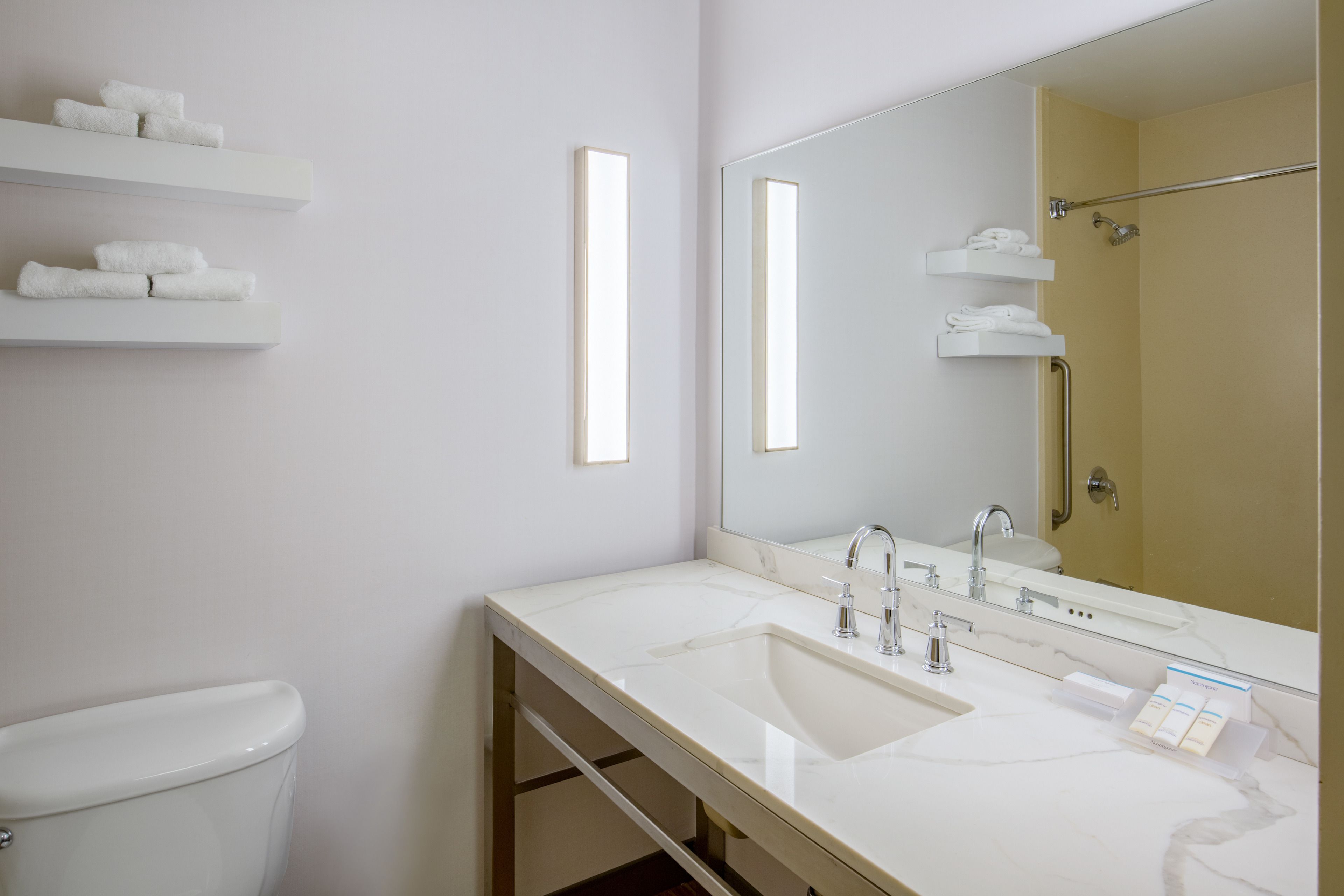 A modern bathroom with a white sink, mirror, and toilet. There are shelves with towels and a shower area visible.