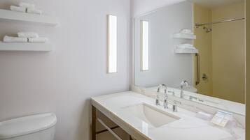 A modern bathroom with a white sink, mirror, and toilet. There are shelves with towels and a shower area visible.