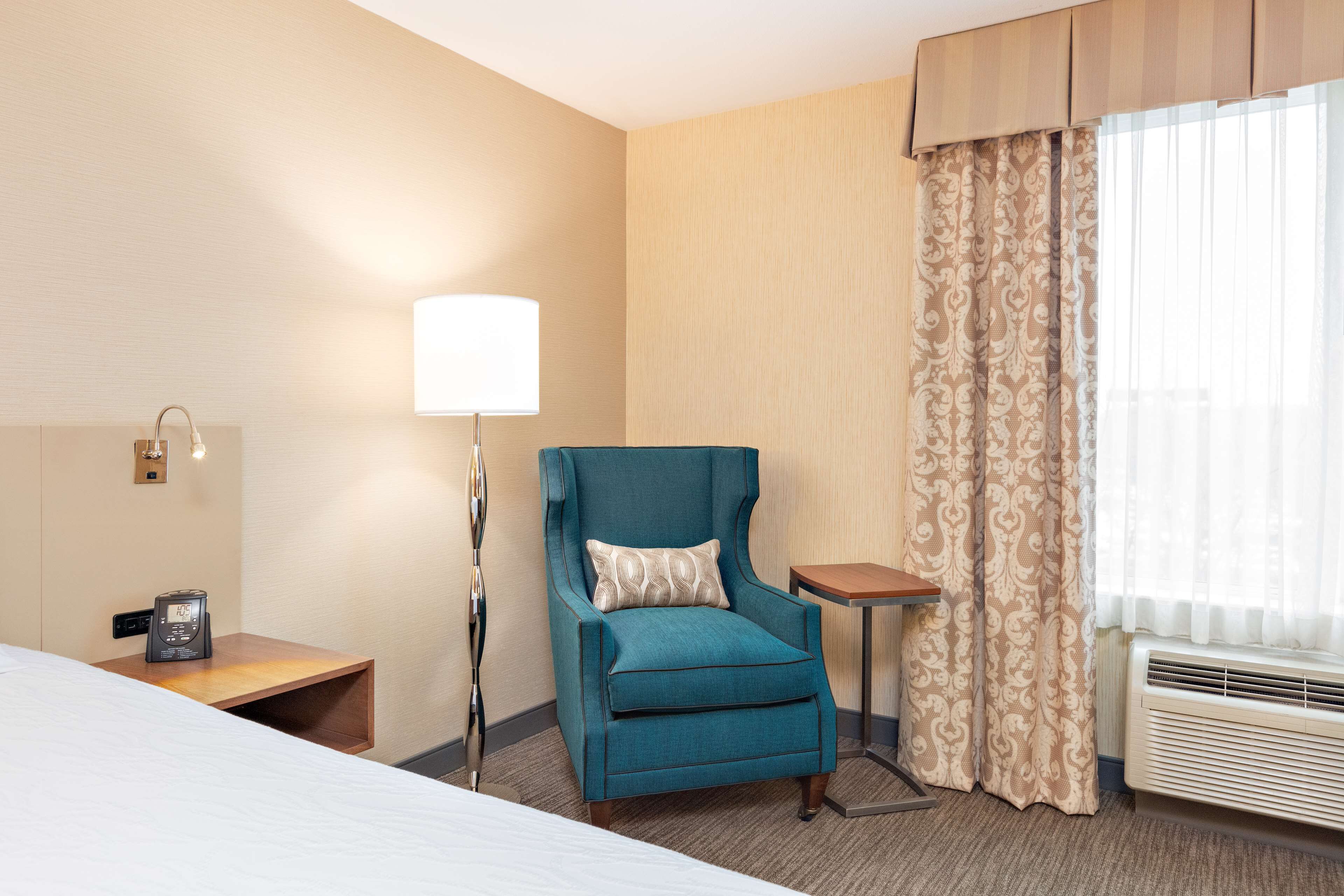 A hotel room with a bed, a teal armchair with a patterned pillow, a bedside table with a lamp, a small table, and a window with curtains.
