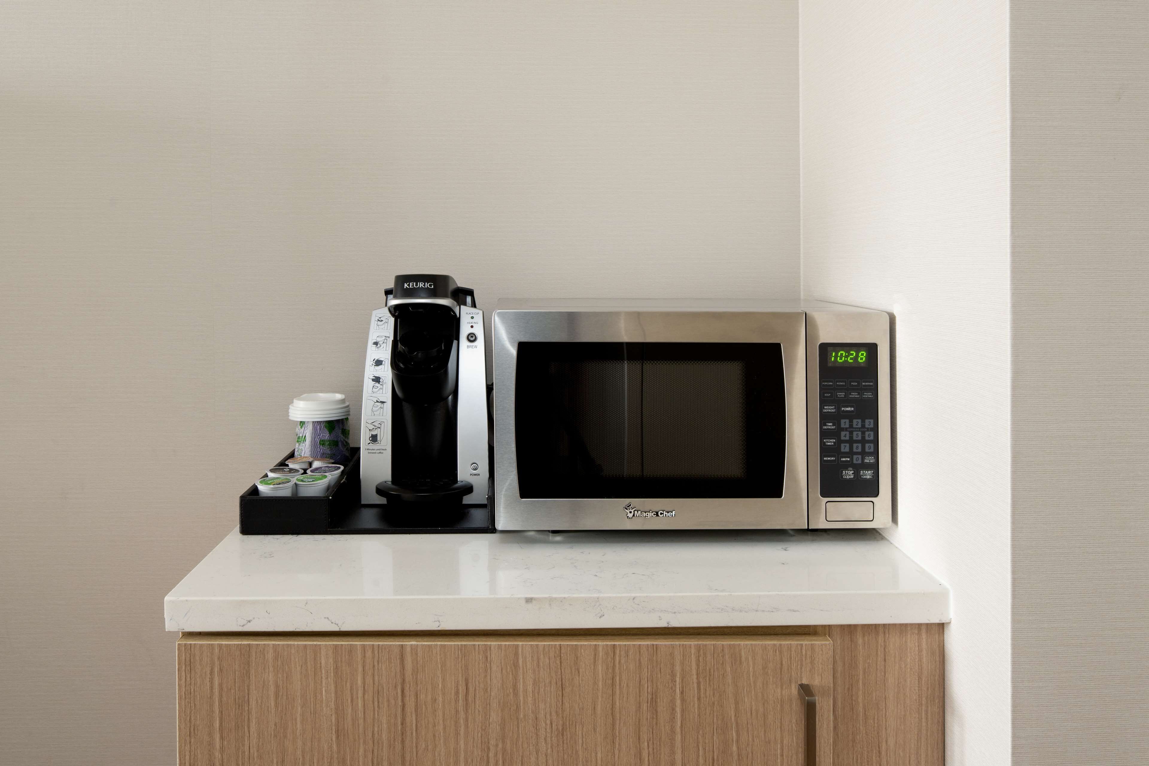 A microwave oven with a digital clock displaying 10:28, a coffee maker, and a small container with coffee pods on a white countertop.