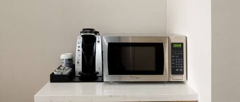 A microwave oven with a digital clock displaying 10:28, a coffee maker, and a small container with coffee pods on a white countertop.