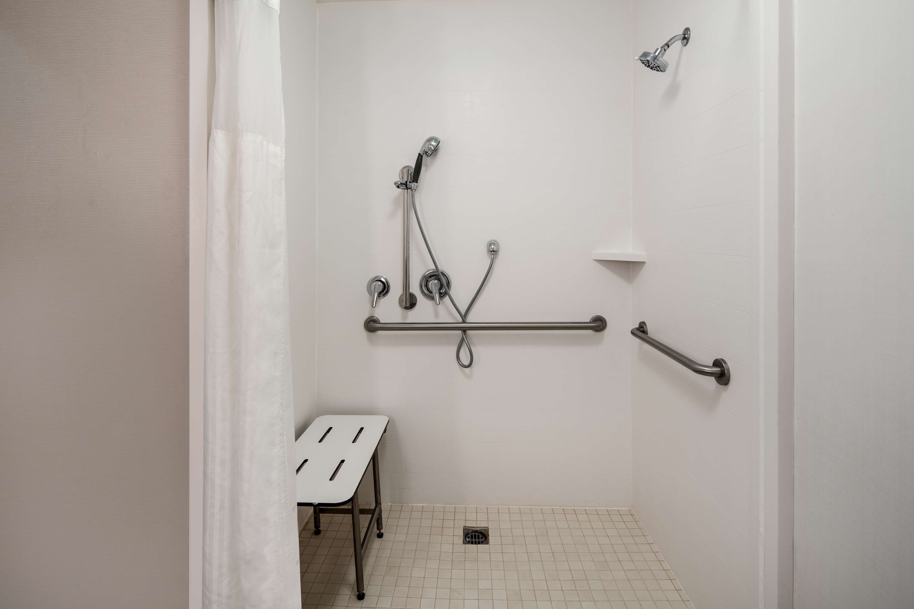A small, white-walled bathroom with a shower, grab bars, and a white bench.