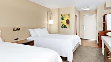 A hotel room with two beds, a bedside table, a lamp, a painting, a door, and a bathroom area.