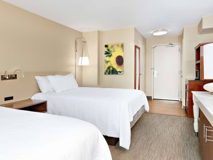 A hotel room with two beds, a bedside table, a lamp, a painting, a door, and a bathroom area.