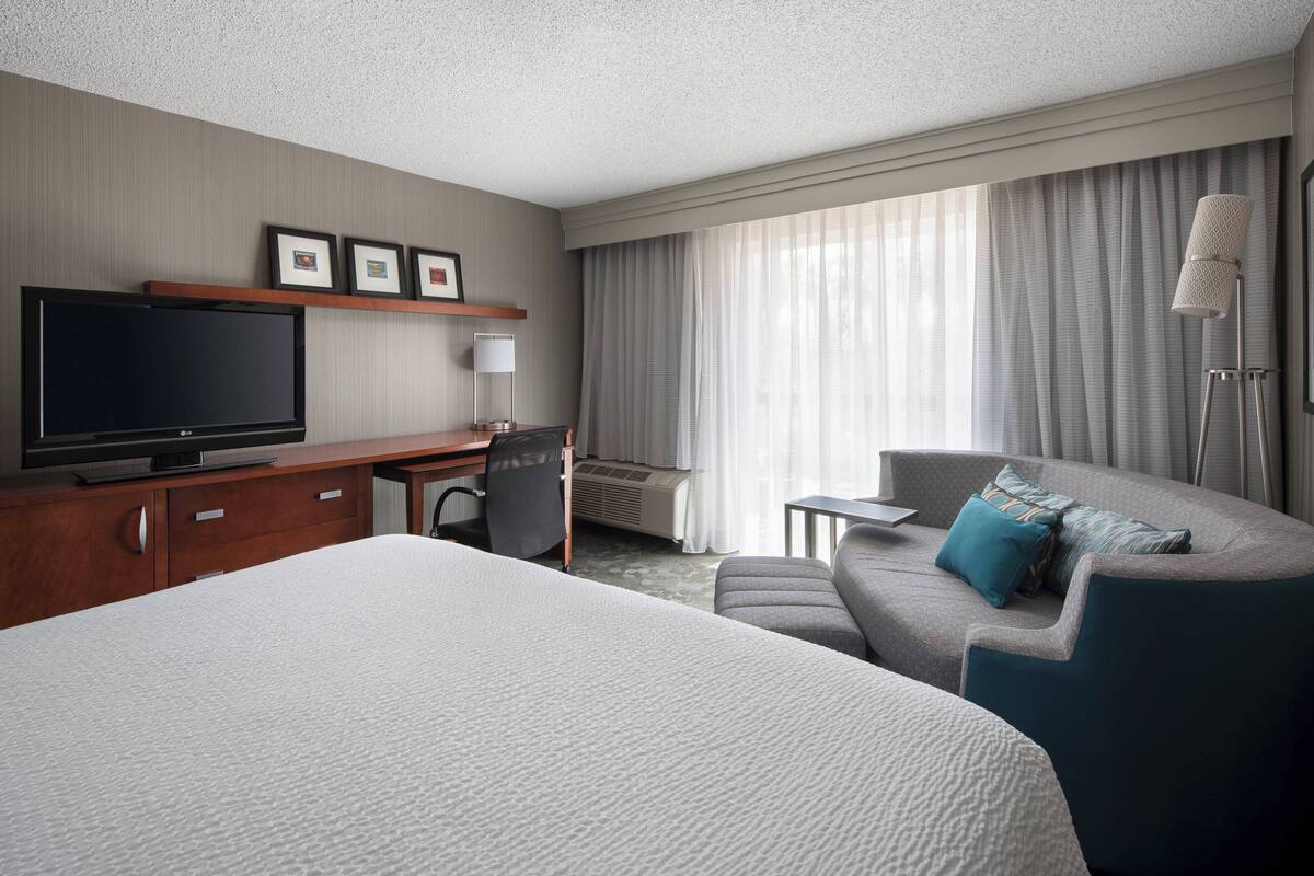 A hotel room with a large bed, a desk with a flat-screen TV, a sofa with pillows, a standing lamp, and a window with curtains.