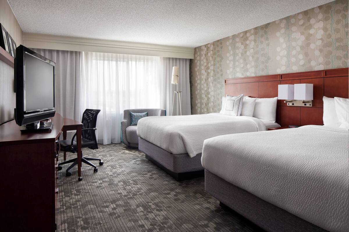 A hotel room with two beds, a desk with a TV, a chair, and a lamp.
