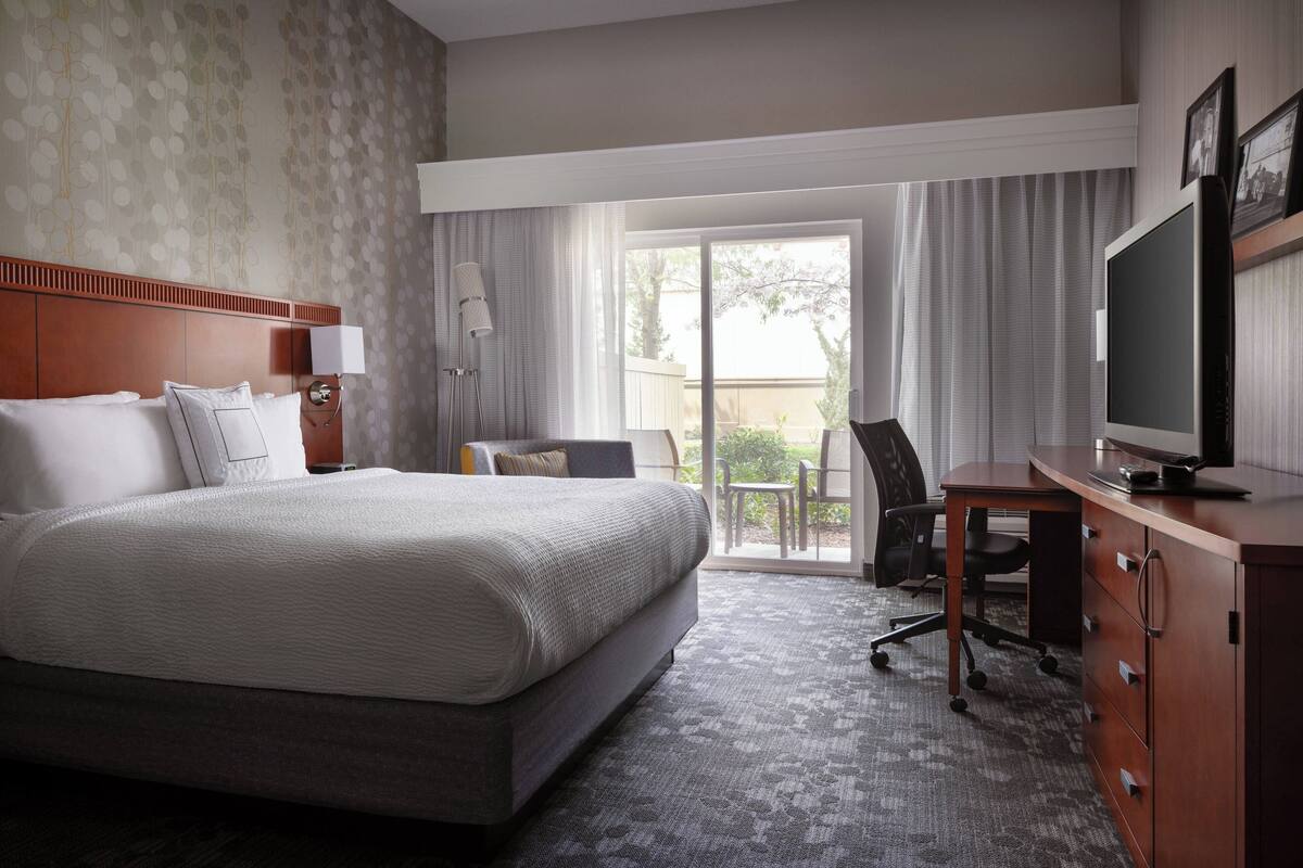 A hotel room with a large bed, a desk with a television, a chair, and a view of the outdoors through a sliding door.