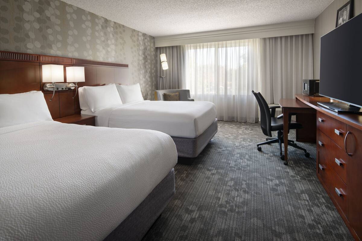 A hotel room with two beds, a desk, a chair, a TV, and a lamp.