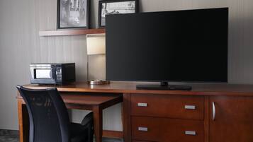 Habitación de hotel con un escritorio de madera, una silla negra, un televisor de pantalla plana, un microondas y un cuadro en la pared.