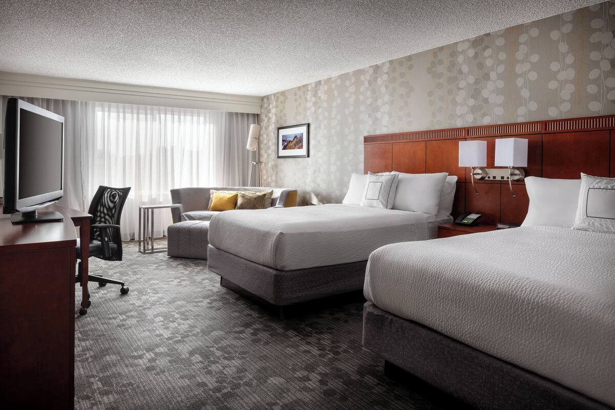 A hotel room with two beds, a desk with a TV, a chair, and a lamp.