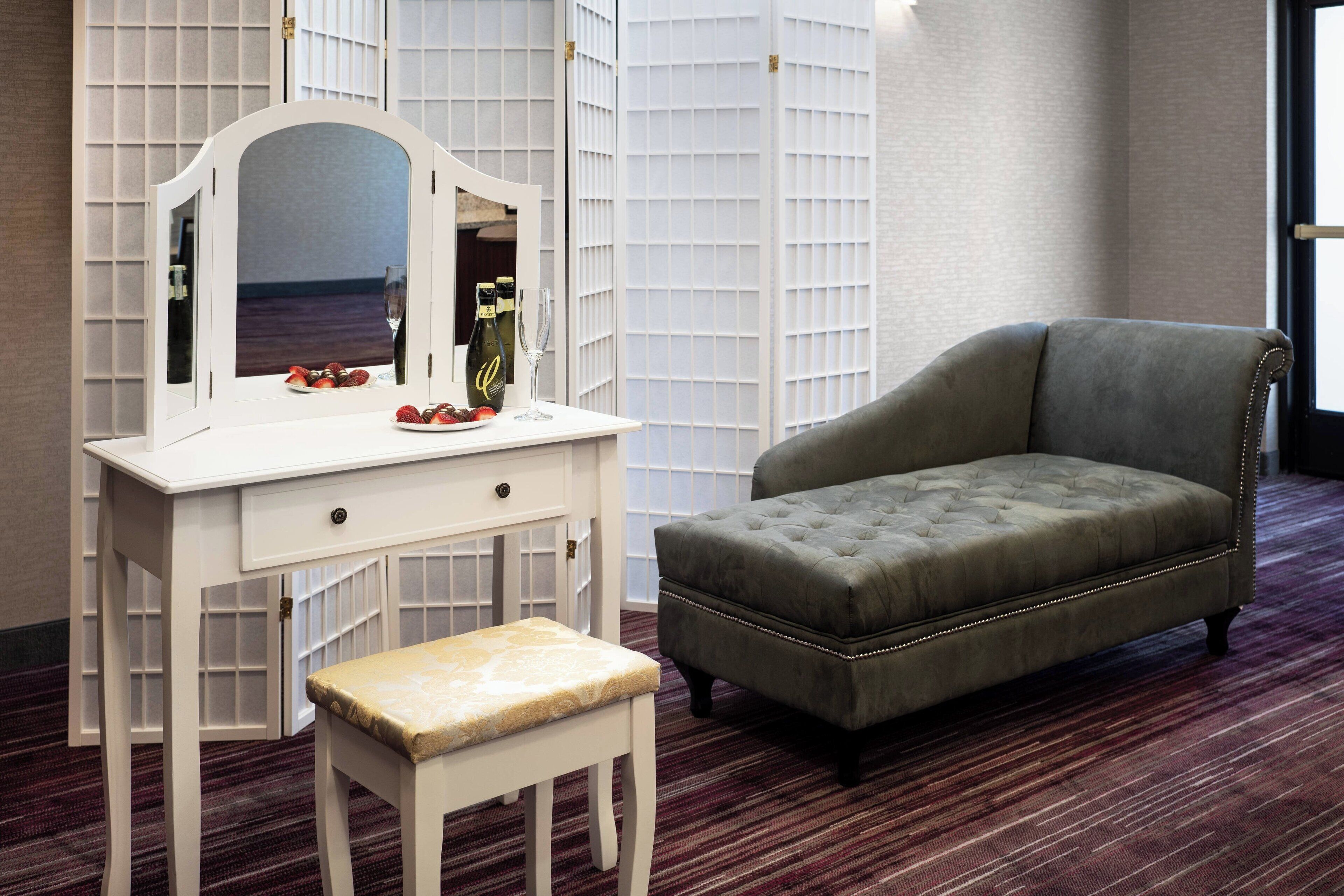 Een kamer met een witte kaptafel, een fauteuil met kussens en een klein krukje.