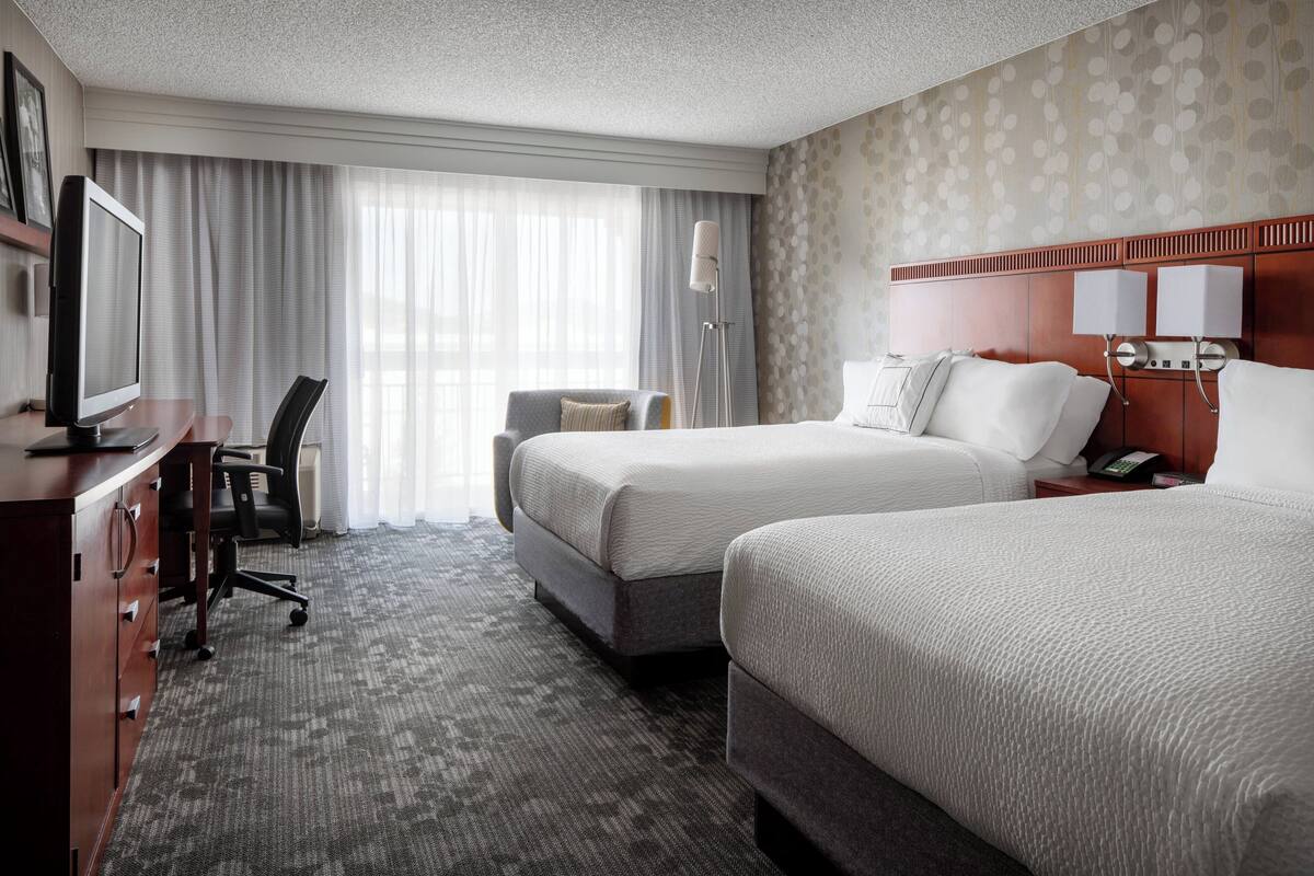 A hotel room with two beds, a desk with a computer, a chair, and a large window with curtains.