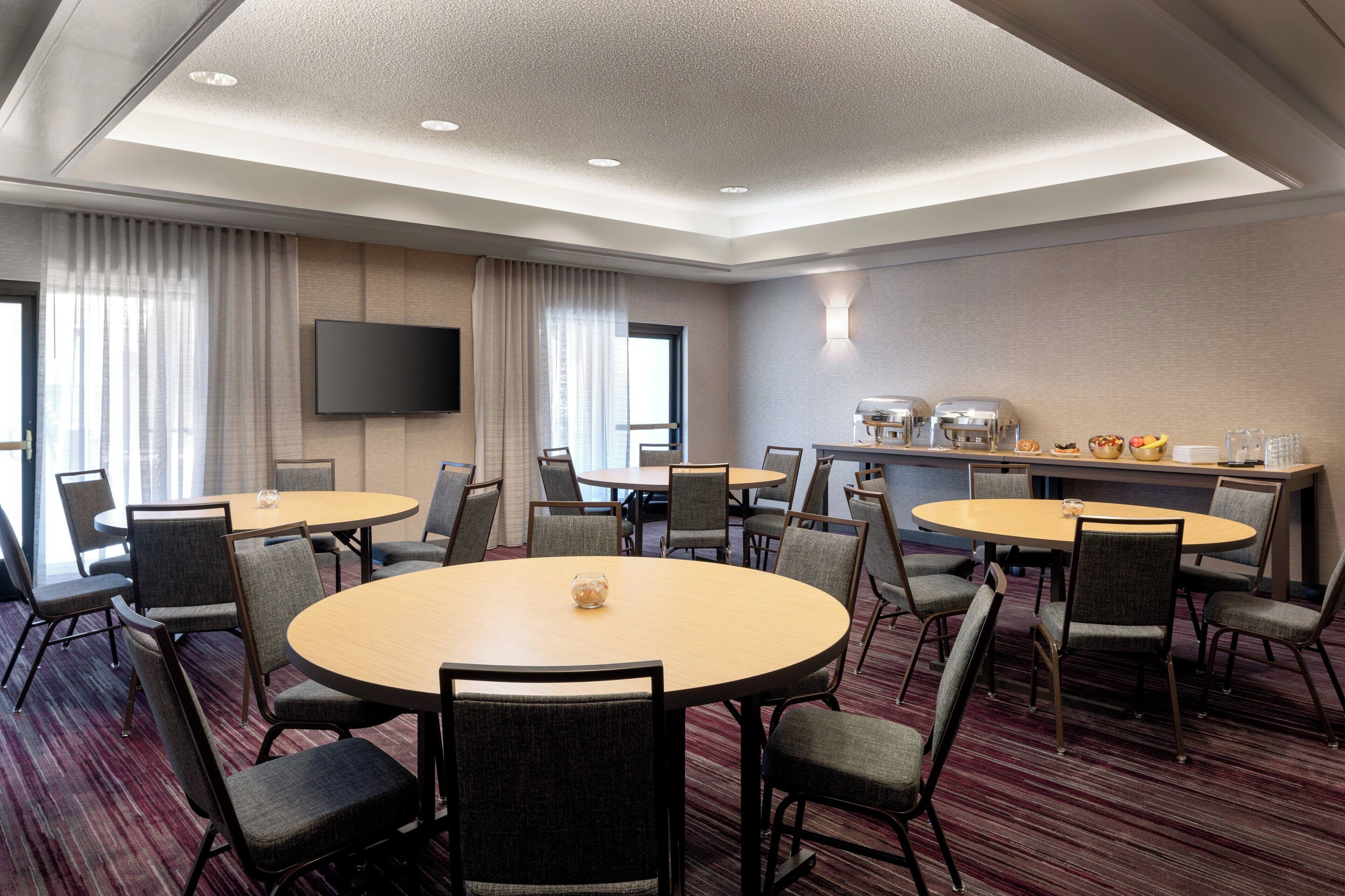 A conference room with round tables and chairs, a flat-screen TV, and a buffet area with food and drinks.